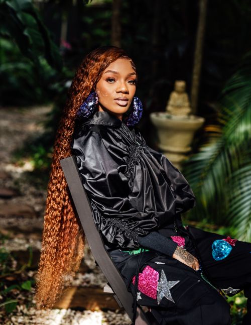 Attractive Black woman with auburn hair and large hoop earrings poses in a black satin jacket with decorative pants outdoors in a lush garden.