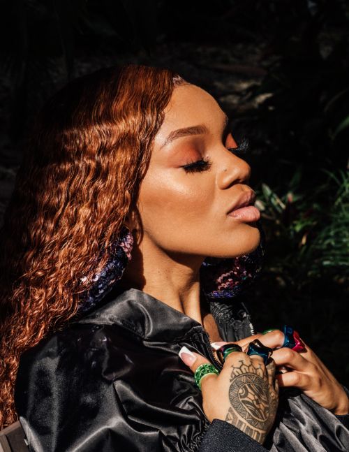 Close-up portrait of a woman with auburn curly hair wearing a black jacket and multiple rings, posing outdoors in natural light.