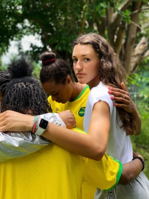 Three female athletes, dressed in Jamaica team attire, embrace outdoors with green trees in the background; focus on their camaraderie.