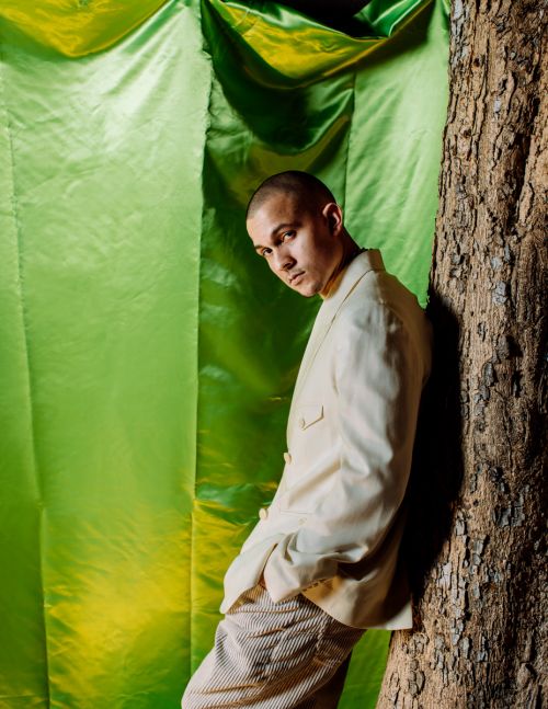 Adult male poses next to a tree in front of a bright green fabric backdrop while wearing a white jacket and striped pants indoors.