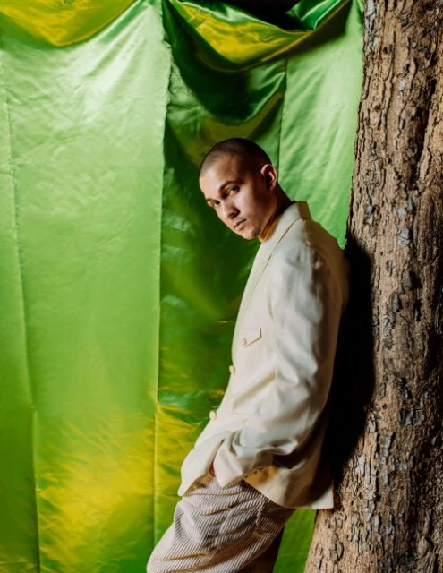 Adult male poses next to a tree in front of a bright green fabric backdrop while wearing a white jacket and striped pants indoors.