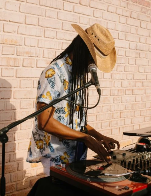 Man with dreadlocks wearing a cowboy hat and floral shirt uses a DJ turntable, with a microphone in front and a brick wall background.