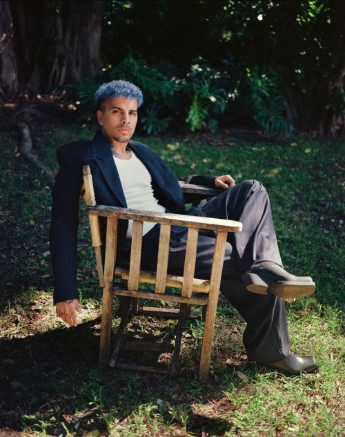 Man with blue hair sits in a wood chair outside in a suit and boots in natural light surrounded by green grass and foliage.