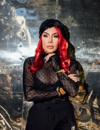 Portrait of a woman with vibrant red hair wearing a black headwrap, sheer monogram top, and dark pants against a reflective metallic wall.