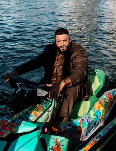 DJ Khaled poses on a green and blue patterned jet ski on a sunny day, wearing a brown suit and watch, set against ocean water.
