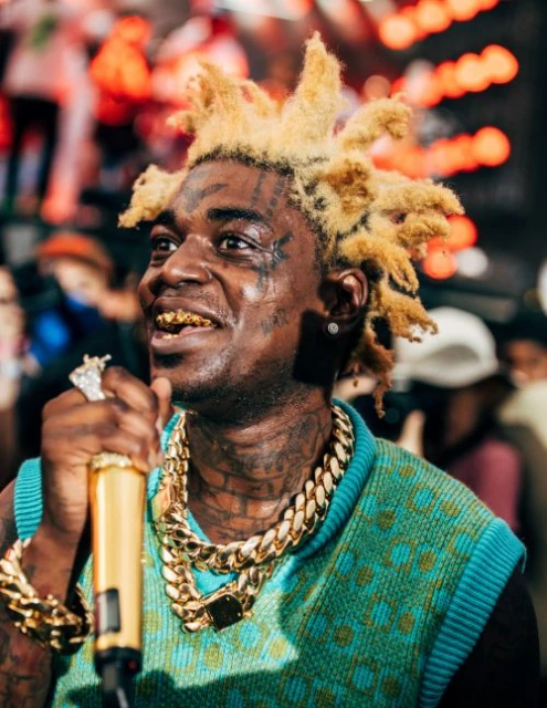 Close-up of a man with blond dreadlocks, gold grill teeth, and heavy gold chains holding a gold microphone while performing.