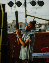 Man with dreadlocks wearing colourful sunglasses singing into a microphone on an outdoor rooftop stage