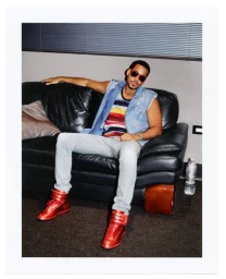 Man wearing sunglasses, denim vest, striped shirt, light jeans, and red high-top sneakers seated on a black leather sofa.