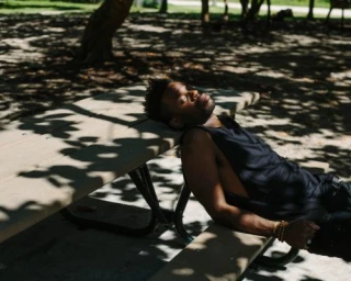 Black man in a black tank top resting on a picnic table bench outdoors under dappled tree shade