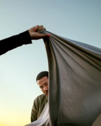 Man peeking from behind a grey fabric sheet held up by a hand against a clear sky background