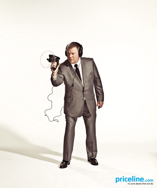 William Shatner holds a vintage movie camera and wears headphones against a bright white background, in a portrait studio shot.