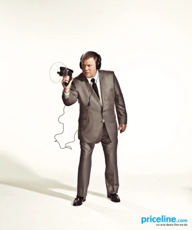 William Shatner holds a vintage movie camera and wears headphones against a bright white background, in a portrait studio shot.