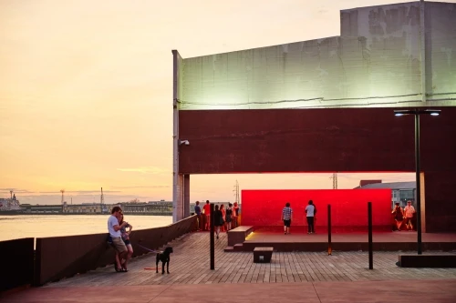 People gather at an outdoor installation with a red wall along a waterfront with a dog at sunset on a wooden platform.