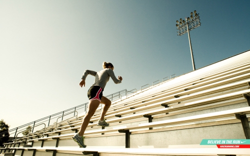 Female athlete runs up the stadium steps in sportswear for exercise, viewed from a low angle on a sunny day with a stadium light.