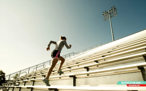 Female athlete runs up the stadium steps in sportswear for exercise, viewed from a low angle on a sunny day with a stadium light.