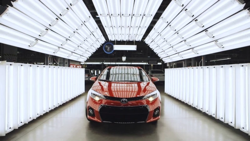 Glossy red car parked inside a brightly lit manufacturing inspection tunnel lined with fluorescent lights