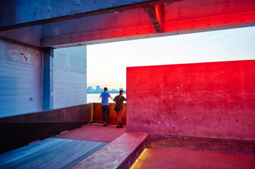 Two people stand together in a concrete structure overlooking water and a distant cityscape, bathed in the glow of dusk lighting.