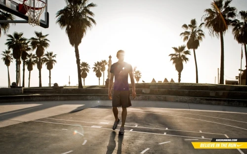 Adult male walking on an outdoor basketball court near palm trees in bright sunlight with a basketball hoop in the background.