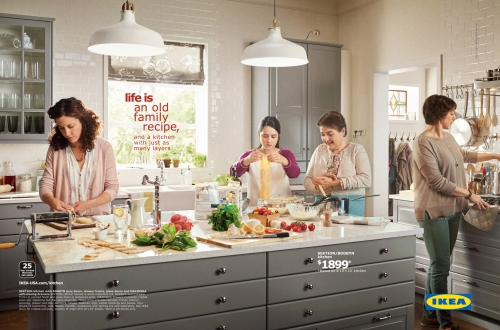 Four women cooking in a gray-toned kitchen with white countertops, pendant lights, and stainless steel appliances. Food prep scene.
