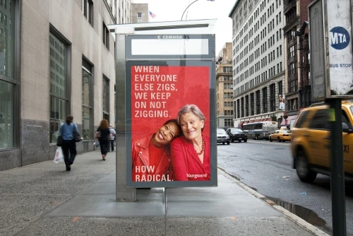 Two women of different ages and ethnicities smile in an advertisement on a bus stop in a city with a yellow taxi passing by.