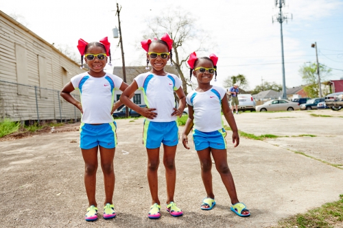 Three smiling African-American girls in matching athletic outfits and red bows stand on a street in front of an old building.