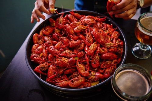 Overhead shot of a large bowl of red crawfish surrounded by people, alongside glasses of beer on a dark surface, great for food themes.