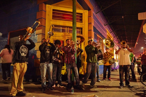 Street band performs on a city sidewalk at night, playing brass instruments beneath the yellow-lit storefront of a building.