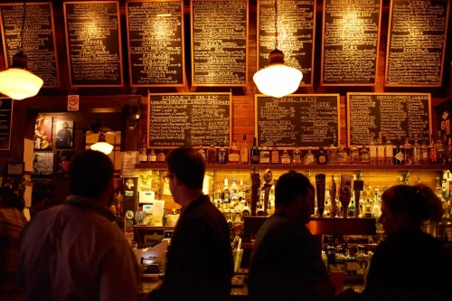 People at a dimly lit bar with wooden decor and chalkboard menus above a wide selection of bottles and drinks on display.