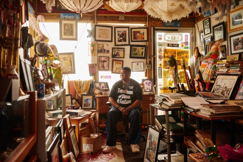 Adult male sits in an interior space surrounded by artwork, framed images, and books in an eclectic and inviting setting.