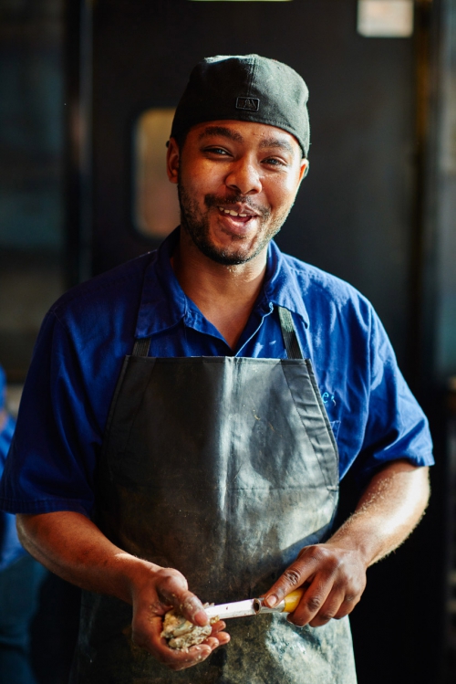 Adult man wearing a black apron and cap is smiling while holding food and a syringe, inside a kitchen with dark background.