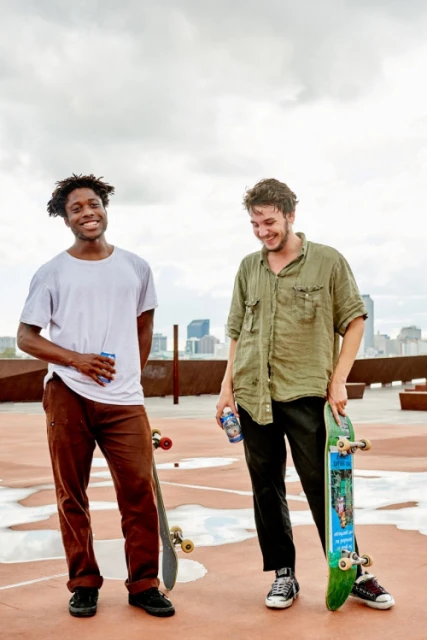 Two young men standing on a rooftop with skateboards, against a cloudy sky and city skyline in the background. Smiling and holding drinks.