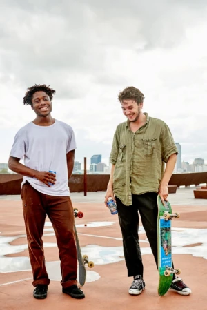 Two young men standing on a rooftop with skateboards, against a cloudy sky and city skyline in the background. Smiling and holding drinks.
