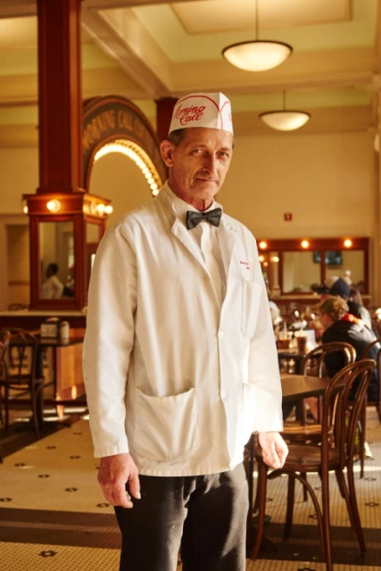 Man in white uniform and hat stands in a restaurant, smiling gently. He is in a vintage-style setting with tables and chairs.