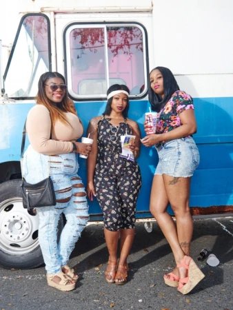 Three fashionable young women pose outdoors in front of a blue and white truck, wearing summer clothes and stylish platform sandals.