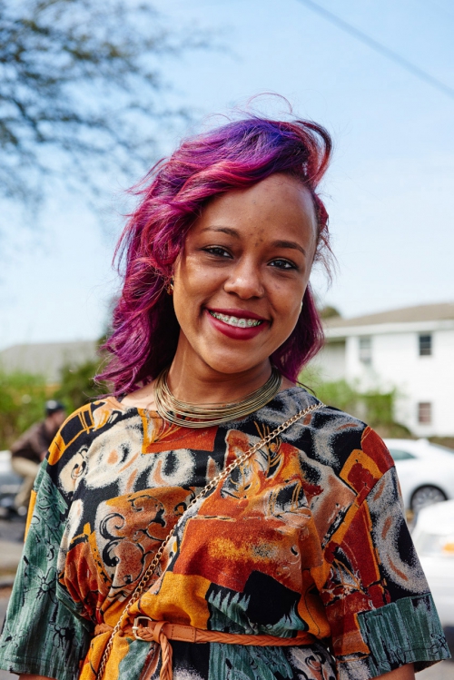 Close-up of a smiling woman with purple hair, wearing a patterned dress and gold necklaces, outdoors with a blue sky background.