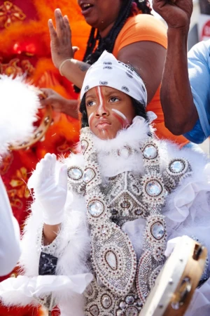 Boy dressed in white beaded and feathered costume and bandanna, wearing face paint and holding drum in outdoor daylight scene.