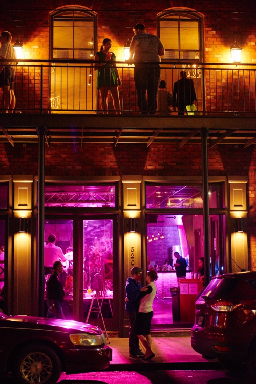 Night scene of a brick building with people on a balcony and a couple embracing in front of a brightly lit bar at night.