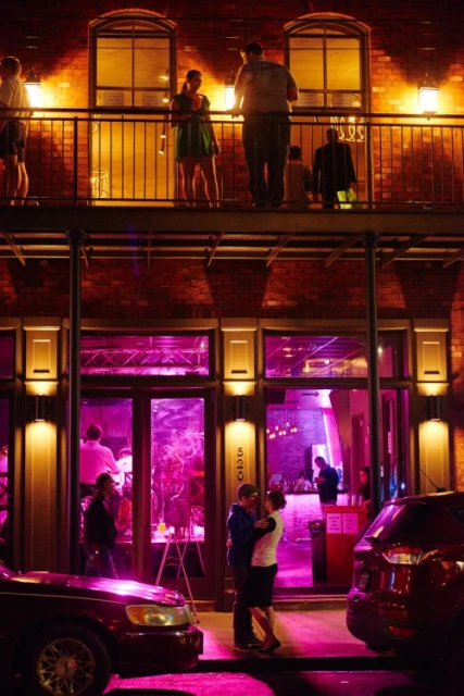Night scene of a brick building with people on a balcony and a couple embracing in front of a brightly lit bar at night.