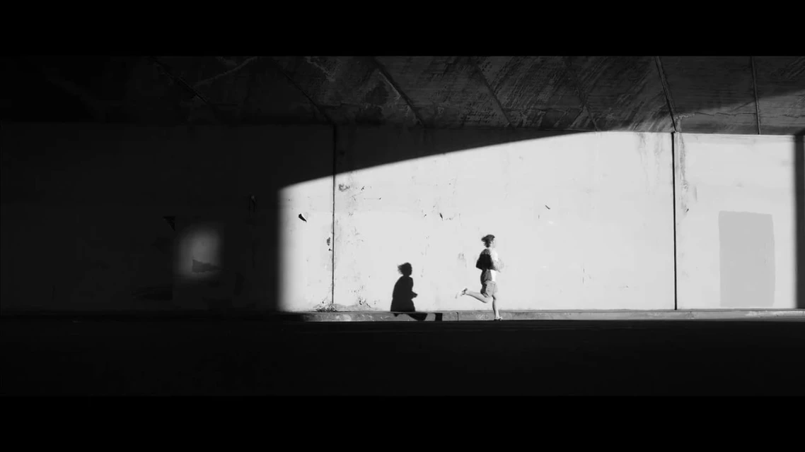 Monochrome image of a person running in an urban underpass, casting a shadow on a sunlit wall, creating a dynamic contrast.