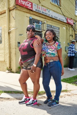 Two African-American women pose outside a yellow brick building with Coca-Cola signs, dressed in casual wear and sneakers on a sunny day.