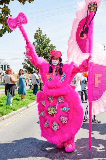 Person in a bright pink costume with face paint marching with a pink hammer and flag during an outdoor parade in daytime.