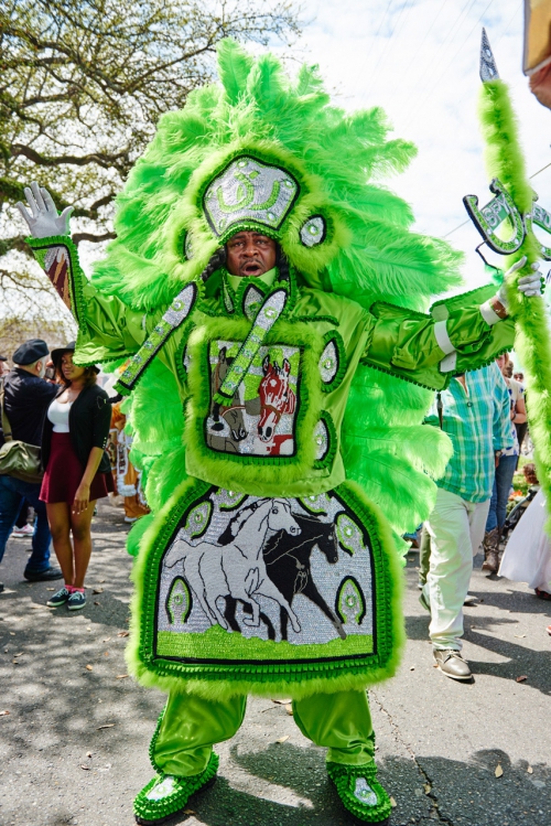 A person in a vibrant, neon green Mardi Gras Indian costume poses outdoors, showcasing intricate beadwork and feather headdress in daylight.
