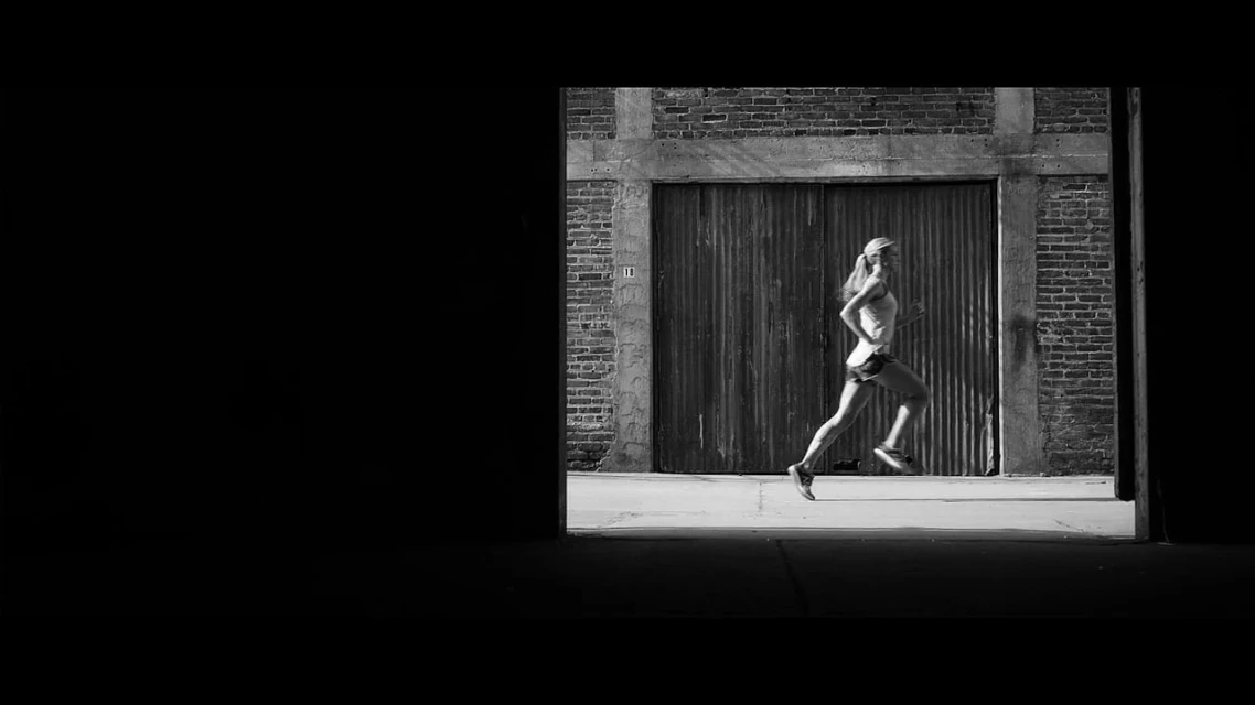 Black and white shot of a woman running on a city sidewalk, framed by a dark tunnel-like opening, with brick building in background.