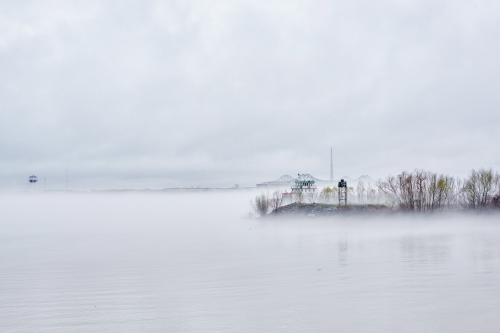 Foggy seascape featuring a distant industrial area, a land bridge, and bare trees, with fog obscuring the sky and water.