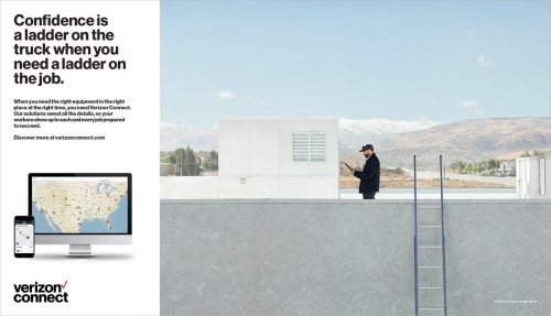 Commercial shot of a man in a jacket and cap standing on a rooftop with a ladder, using a tablet, with mountains in the distance.