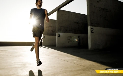 Athletic man running outdoors on concrete surface near concrete walls in bright sunlight, wearing dark athletic wear and running shoes.