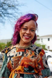 Smiling woman with vibrant purple hair wearing a patterned dress and layered necklace outdoors