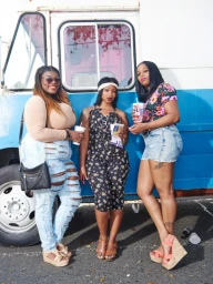 Three women standing outside in front of a parked blue and white van, holding cups.
