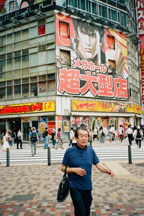 Adult male walks on crosswalk in urban area with commercial buildings and large anime-style advertisement in daytime under overcast lighting.