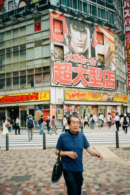 Adult male walks on crosswalk in urban area with commercial buildings and large anime-style advertisement in daytime under overcast lighting.